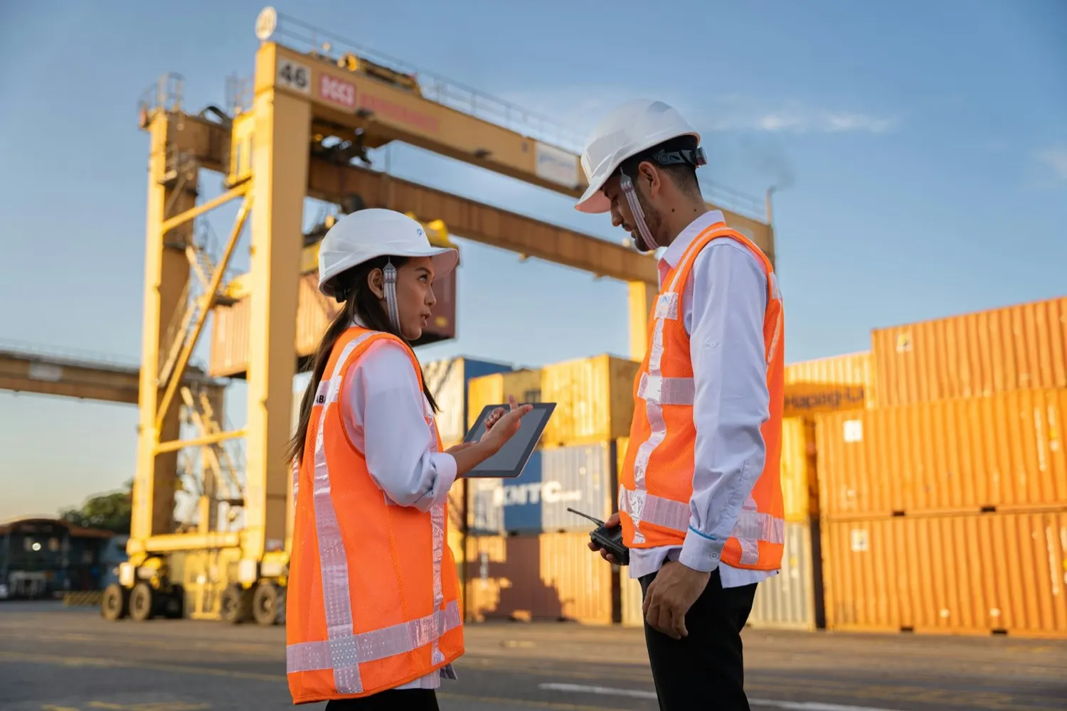 Two workers in high-visibility vests reviewing data on a tablet beside shipping containers at an export terminal